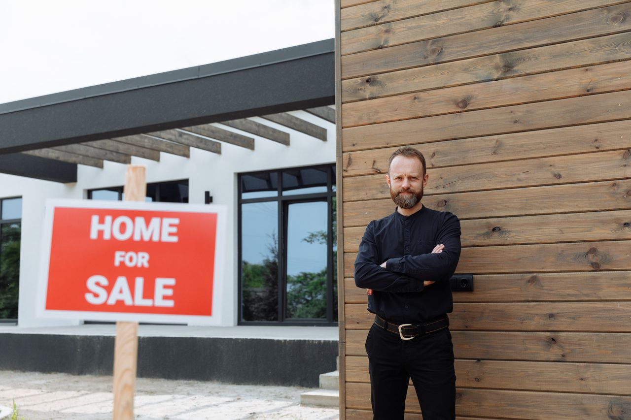 gallery-1 Real estate agent standing by a modern home with a 'Home for Sale' sign.
